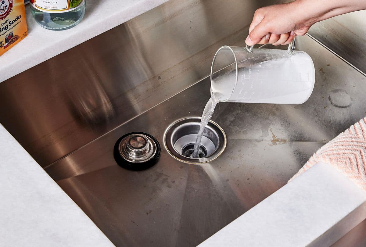Pouring a natural cleaning solution down a kitchen drain to kill drain flies and remove organic buildup.